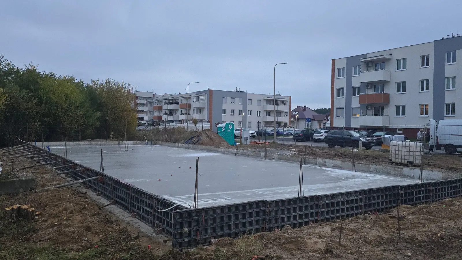 Świeżo zabetonowana płyta fundamentowa w czarnym szalunku systemowym, w tle bloki mieszkalne