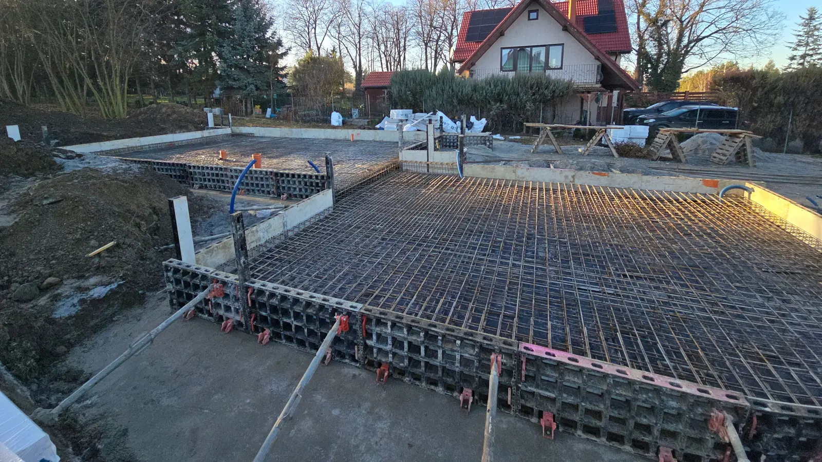 Zbrojenie płyty fundamentowej w szalunku systemowym widziane z narożnika, w tle dom jednorodzinny