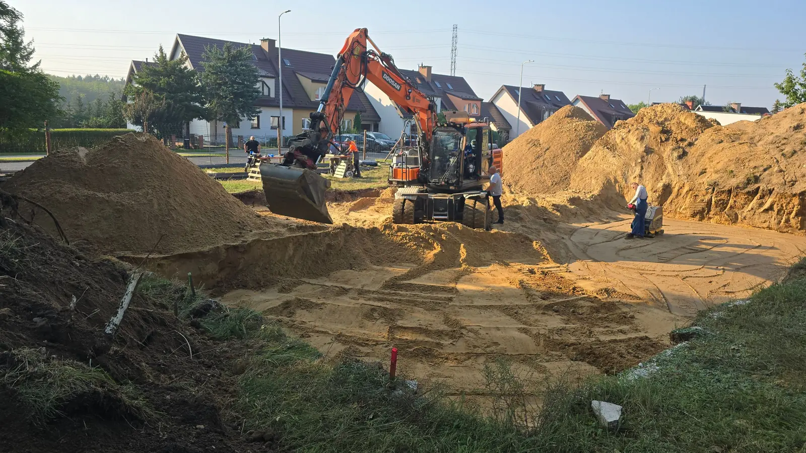 Koparka przy wykopie pod płytę fundamentową, w tle domy jednorodzinne