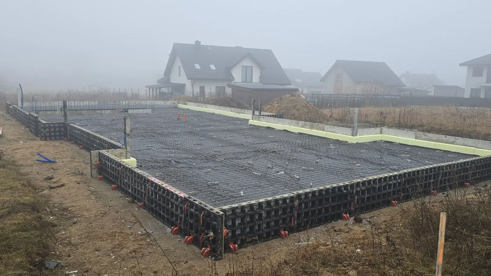Szalunek systemowy z jasnymi kształtkami brzegowymi na obwodzie płyty fundamentowej, w tle domy we mgle